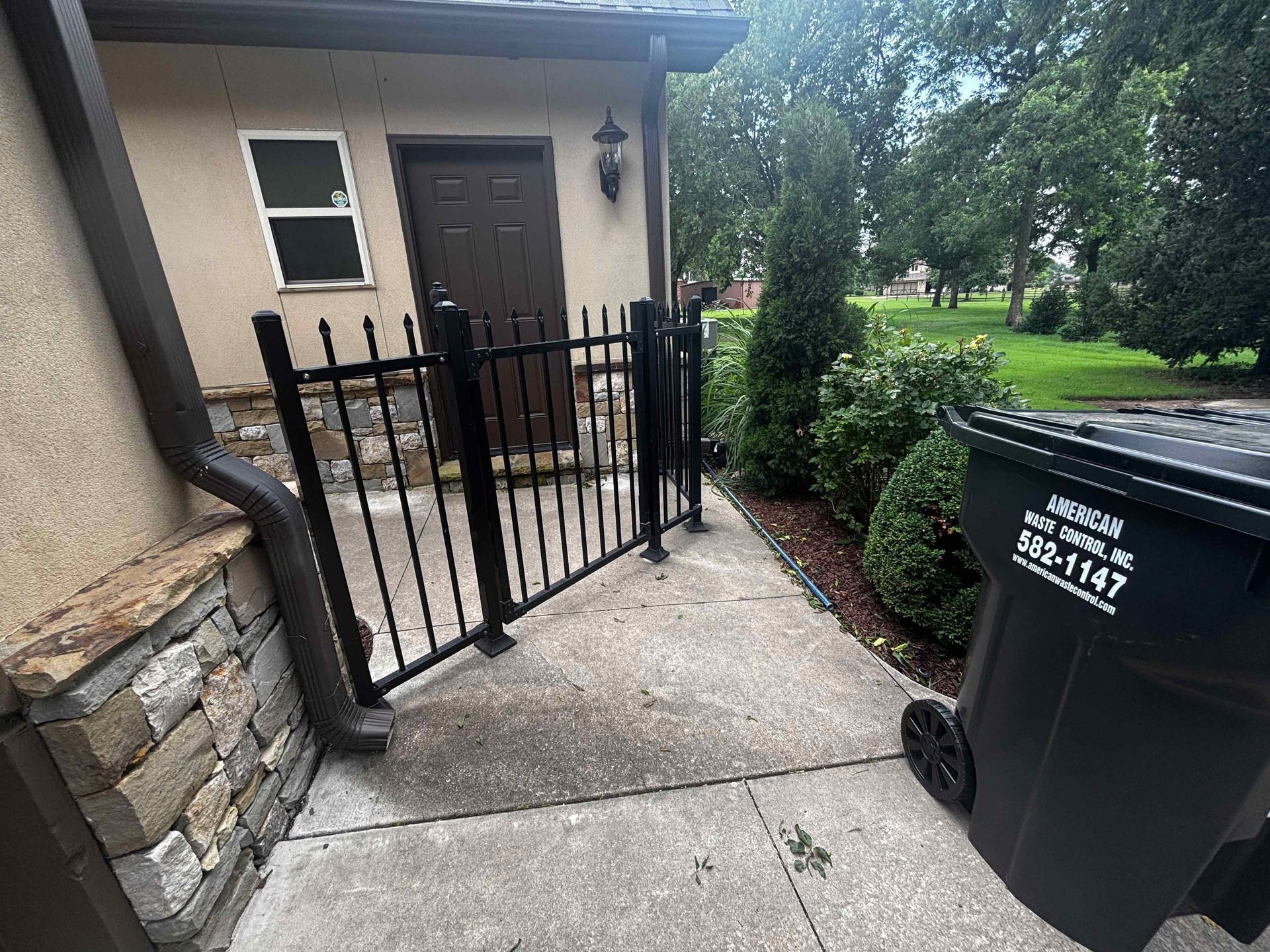 Front walkway to a beige house with a black gate, stone wall, and trash bin beside a green lawn.