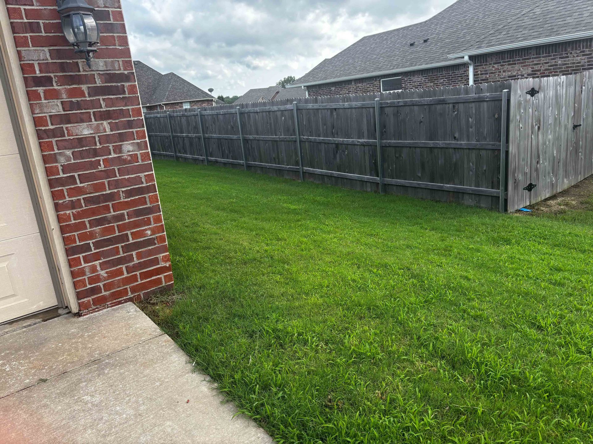 Backyard with green grass, wooden fence, and a brick house corner beside a concrete patio