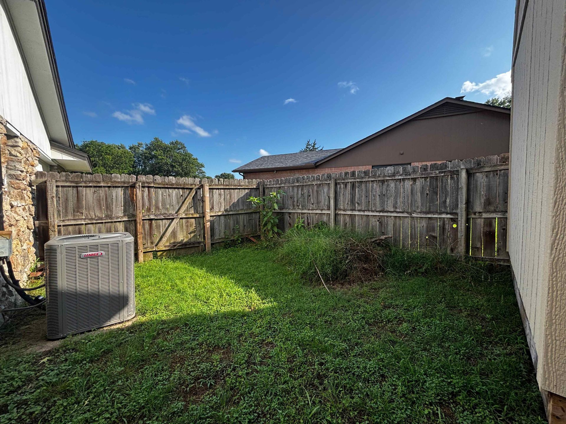 Small fenced backyard with patchy grass, wooden fences, and a gray air-conditioning unit under a blue sky.