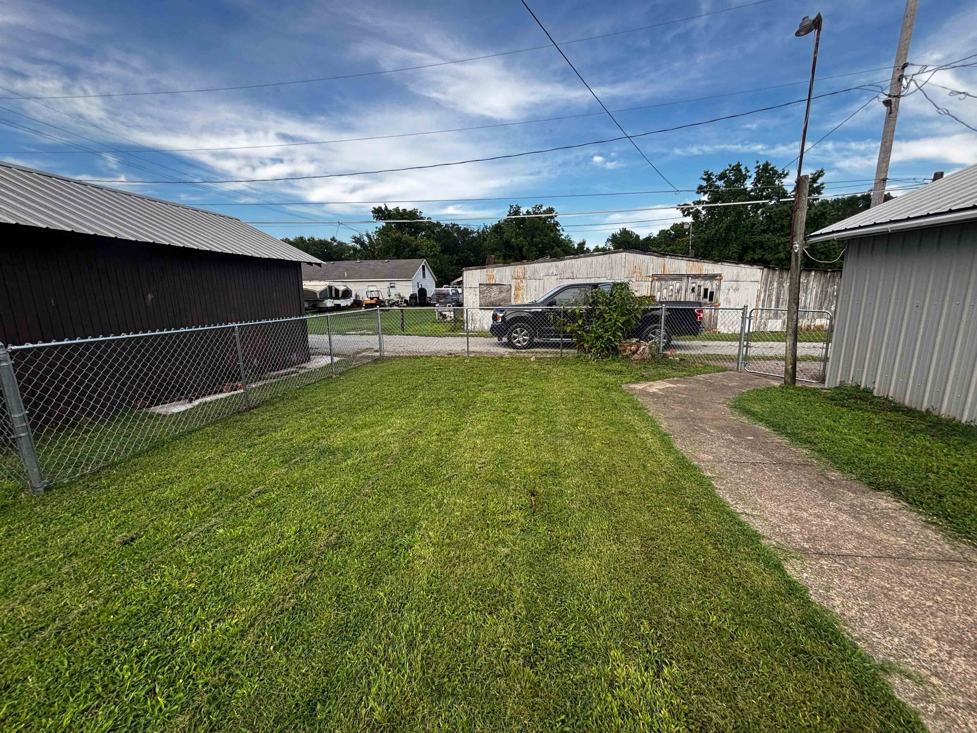 Backyard with green lawn, gravel path, chain-link fence, and sheds under a partly cloudy sky