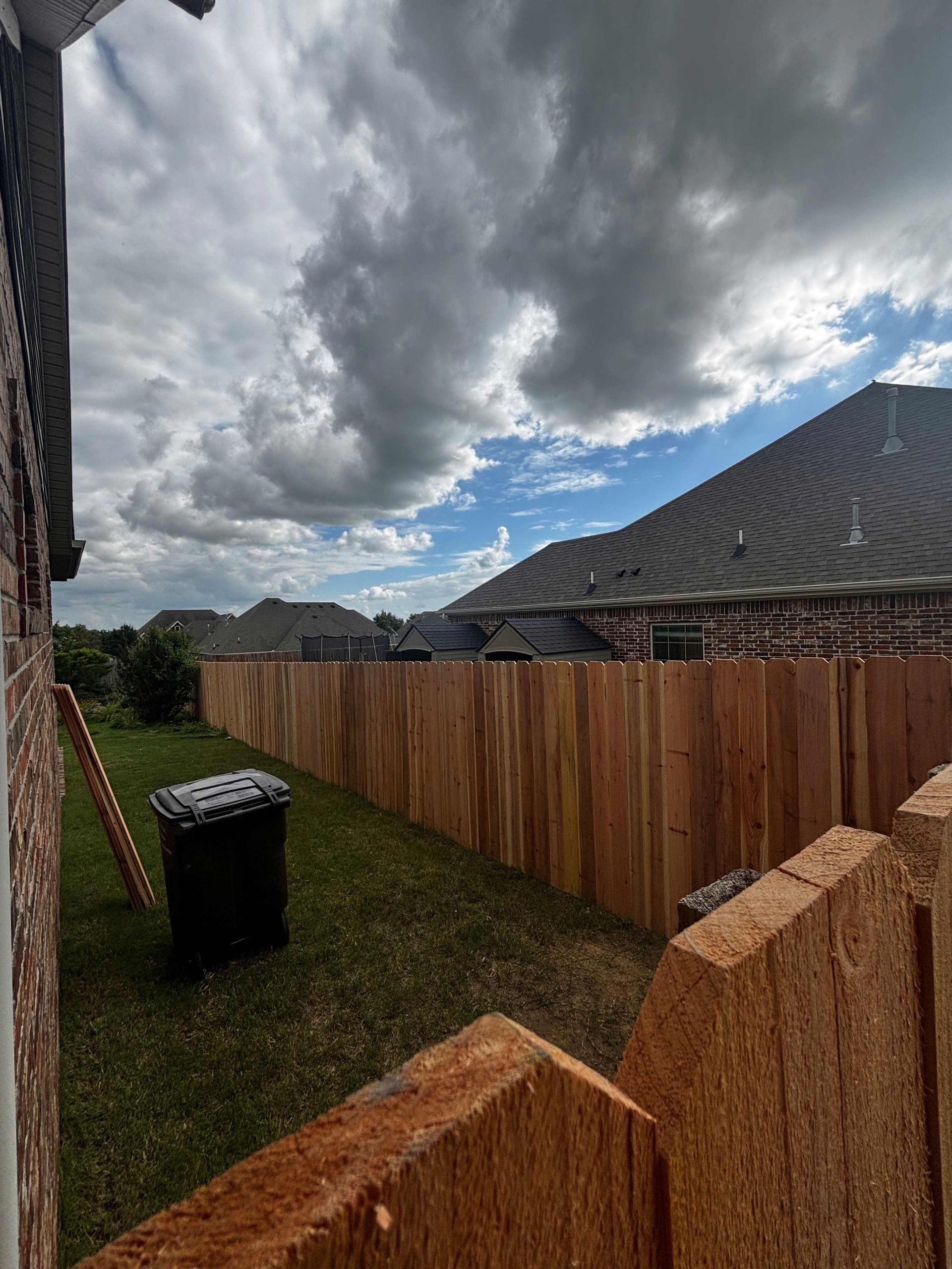 Backyard with wooden fence, black trash bin, and cloudy sky over neighboring houses