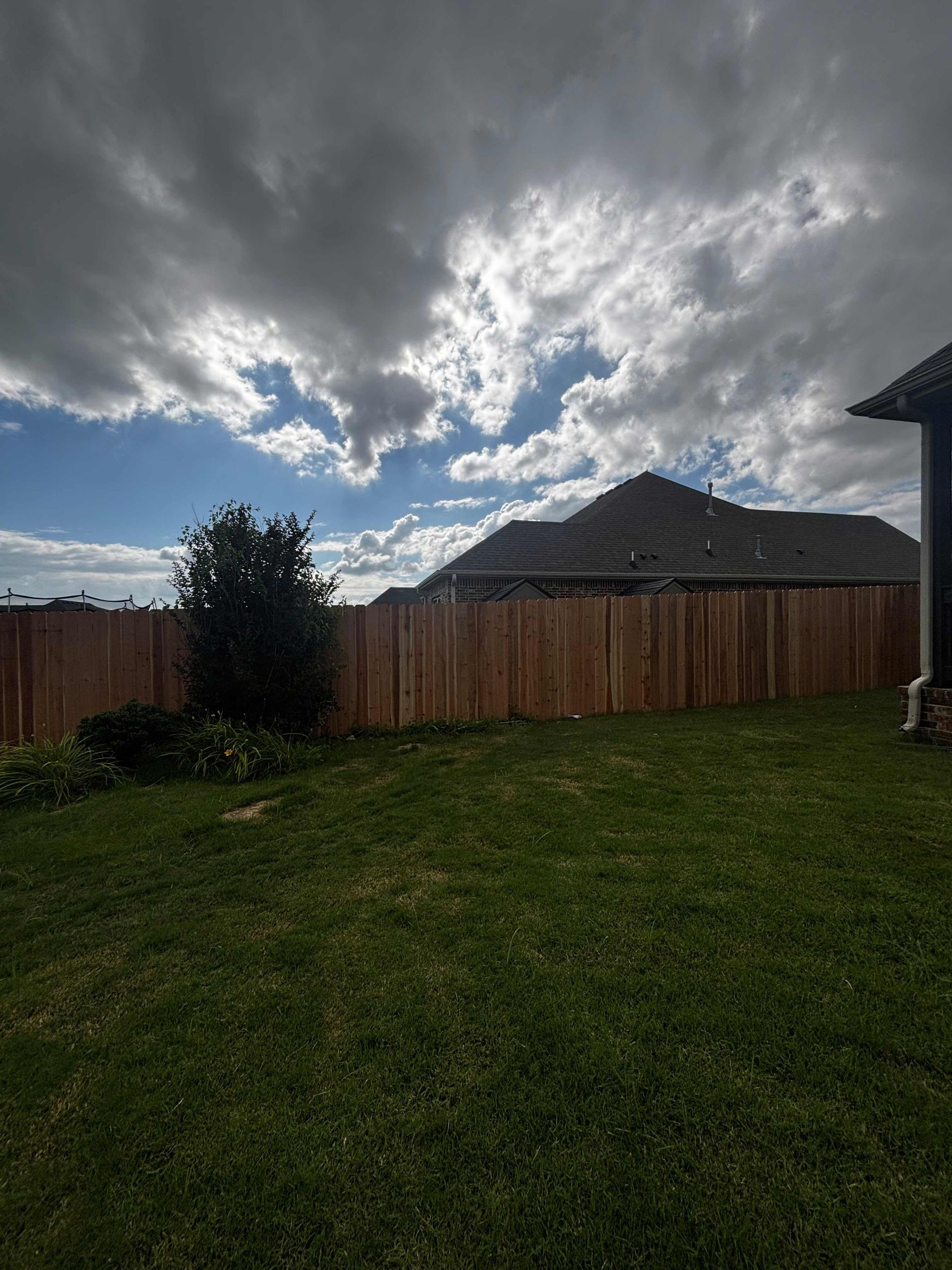Stormy sky over a fenced backyard with a grassy lawn and a house roof in the distance