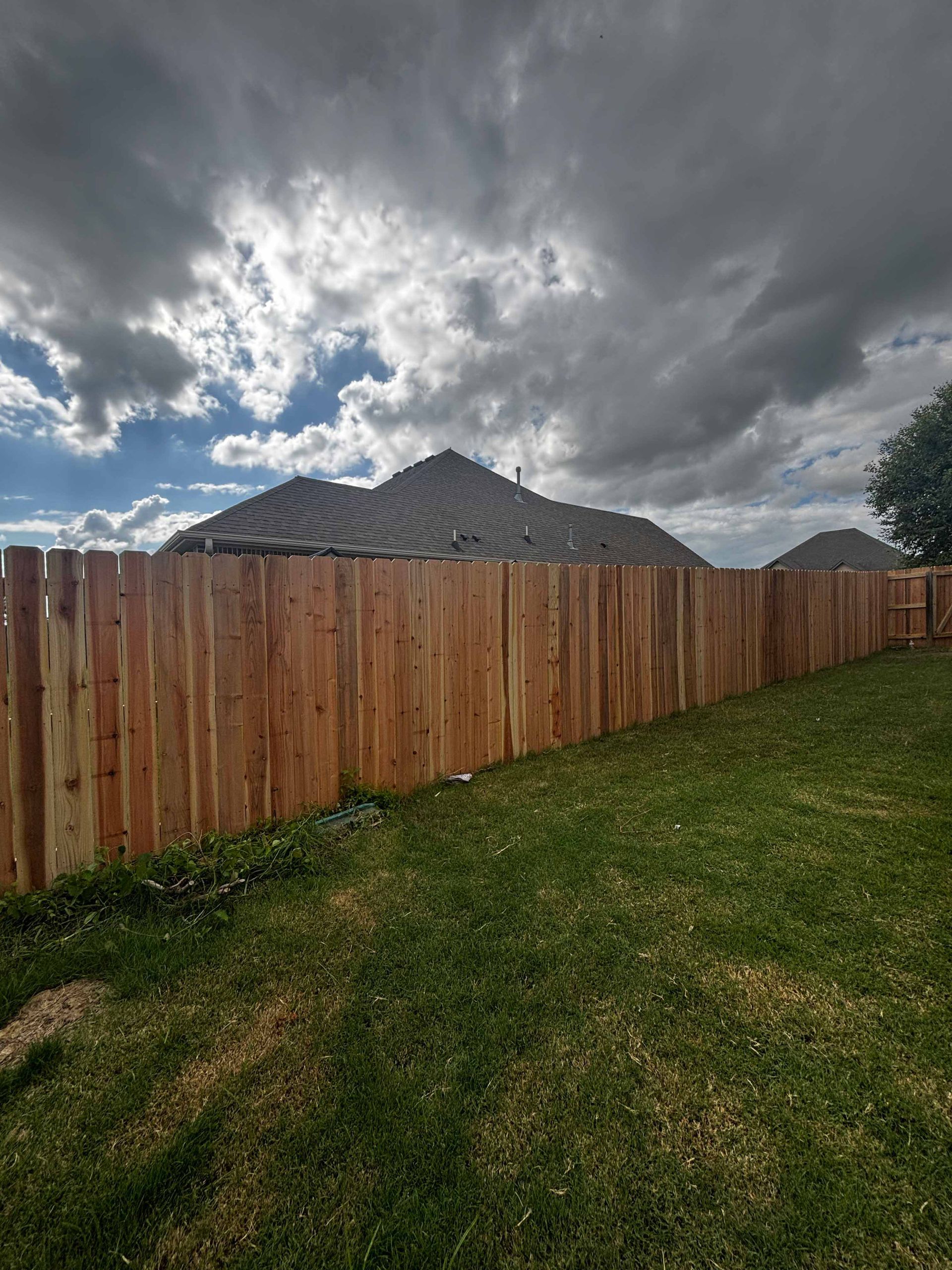 Wooden privacy fence beside a grassy yard under dark storm clouds, with houses in the distance.