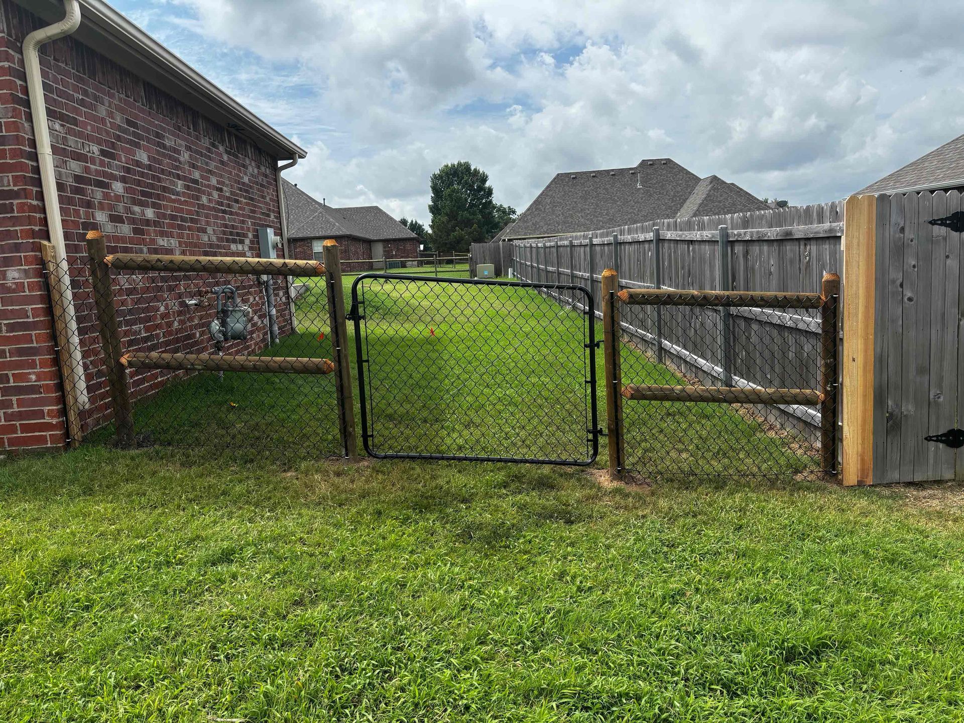 Backyard with open chain-link gate, green grass, and brick house beside a wooden fence under cloudy sky