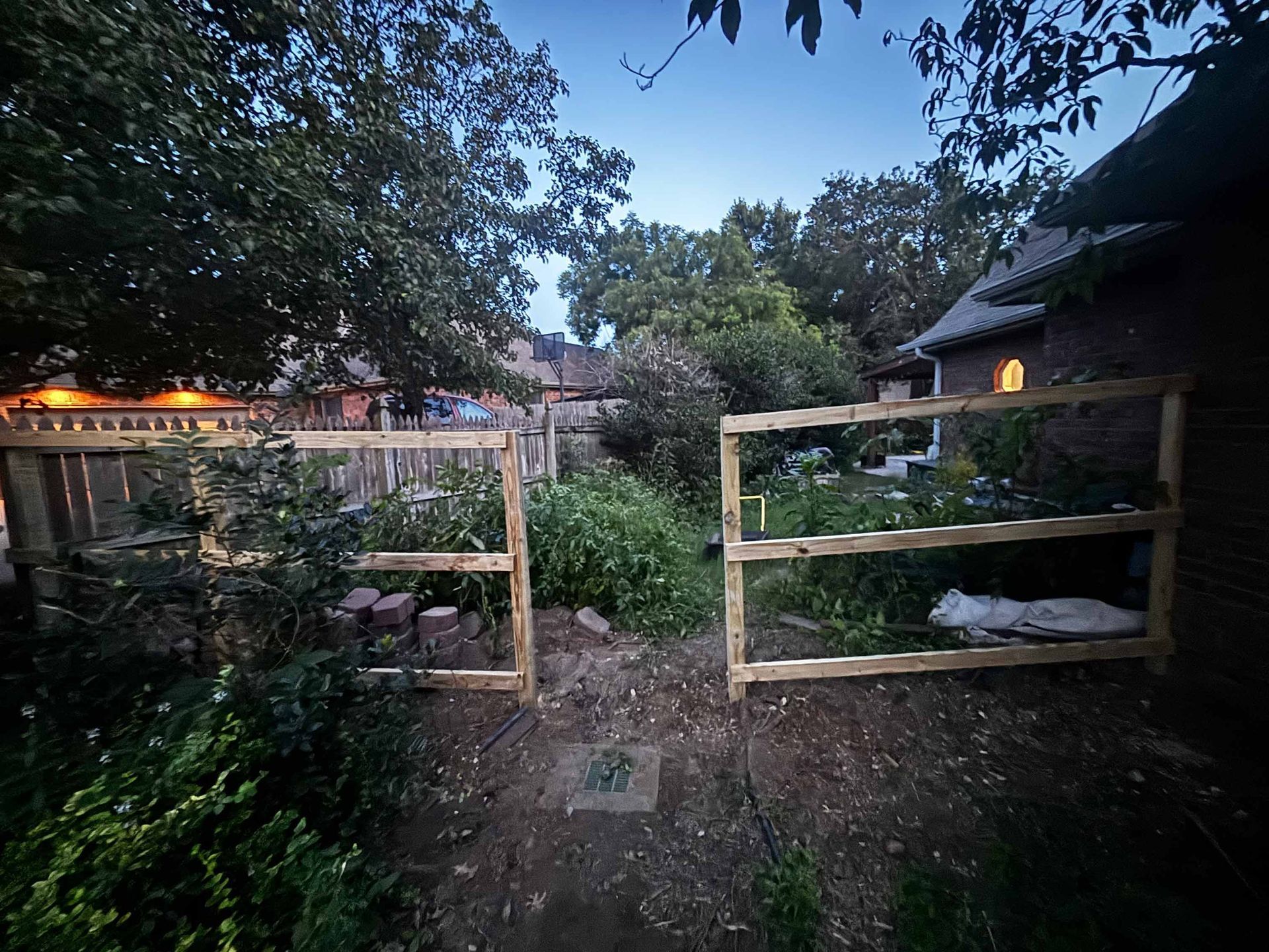 Wooden garden gate at dusk, with a stone path, bushes, and lit houses on both sides.