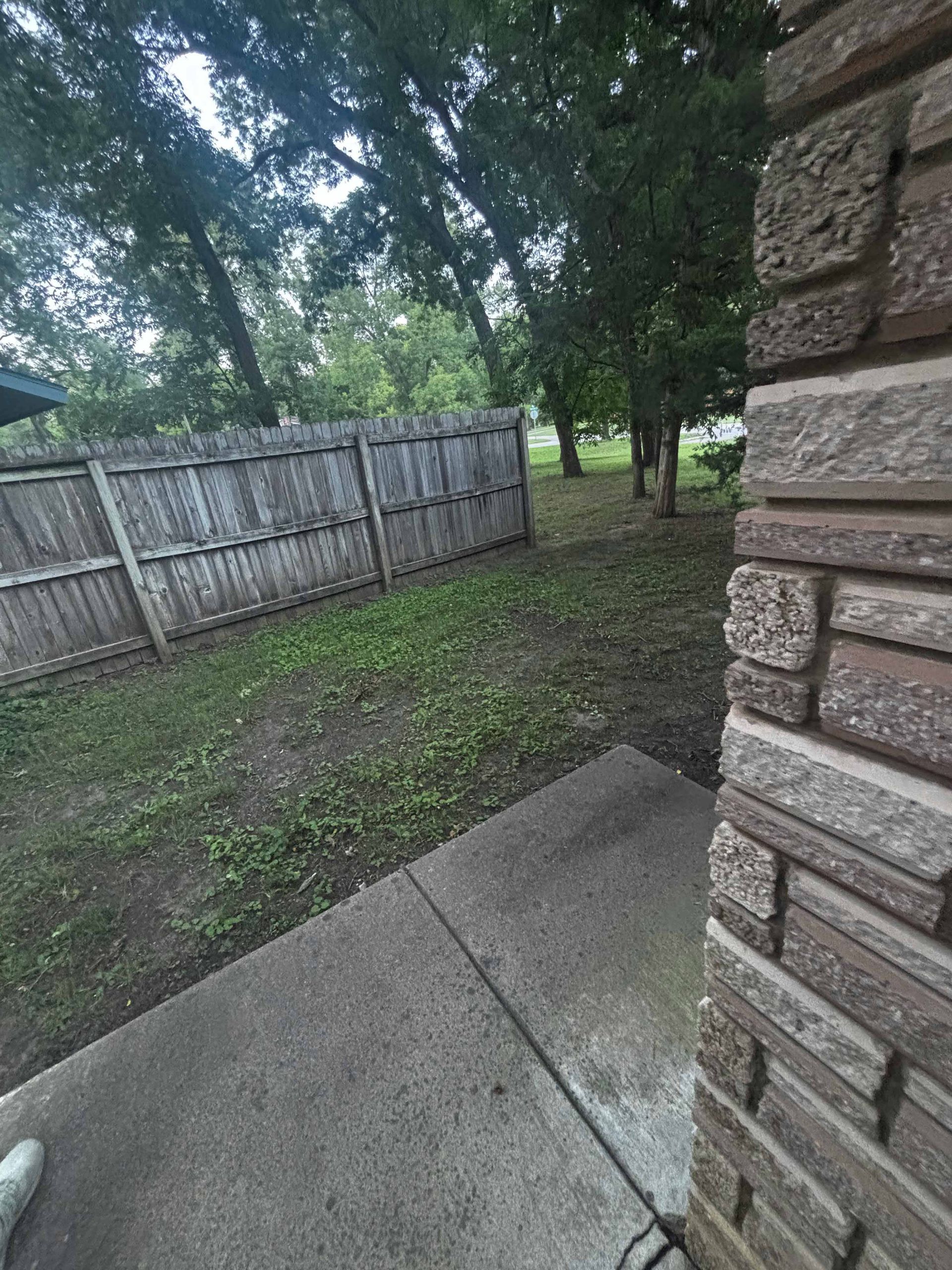 Backyard patio beside a brick house, with a wooden fence and trees in a shaded yard.