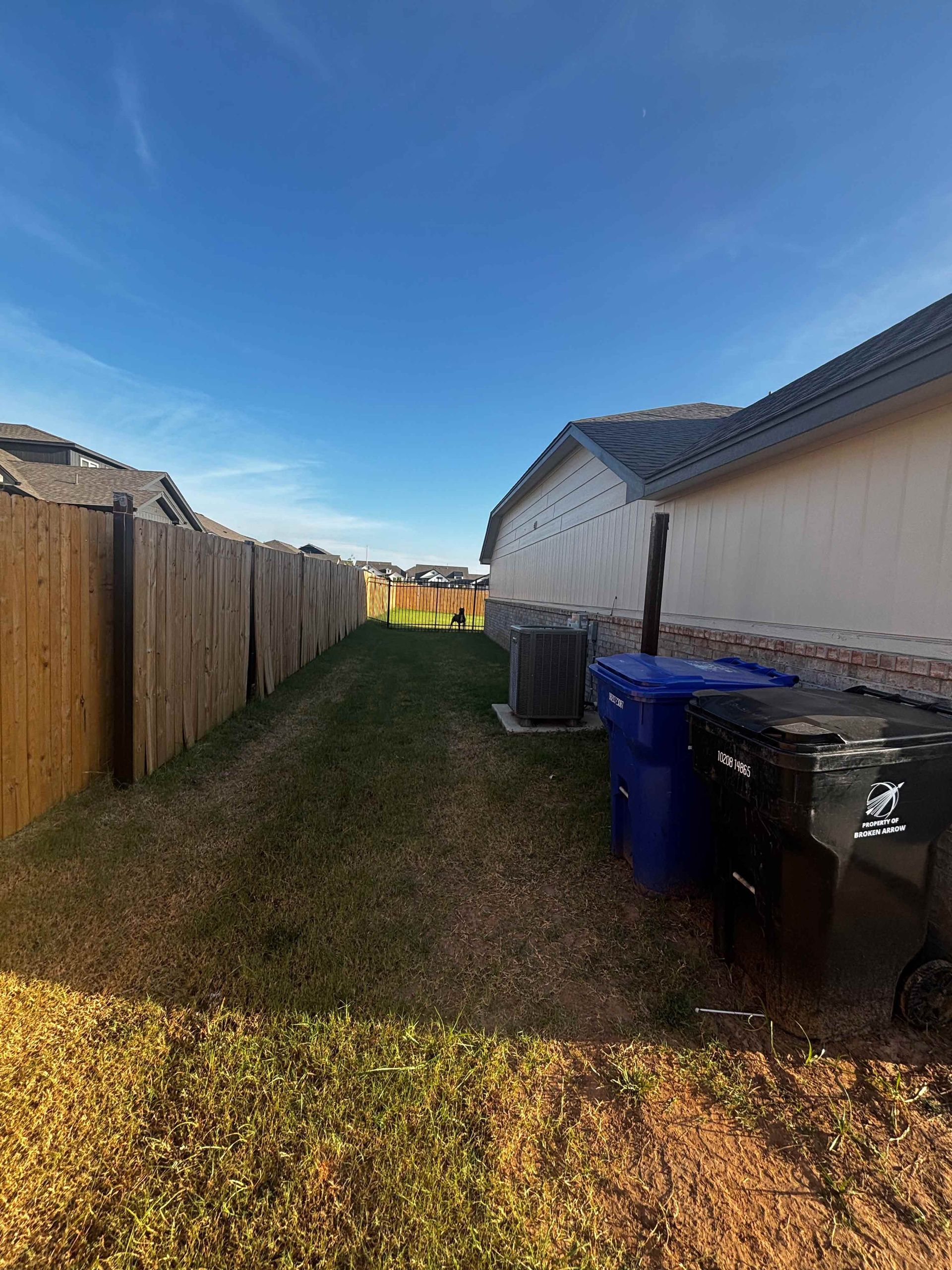 Narrow grassy side yard between a wood fence and white house, with trash bins under a blue sky.