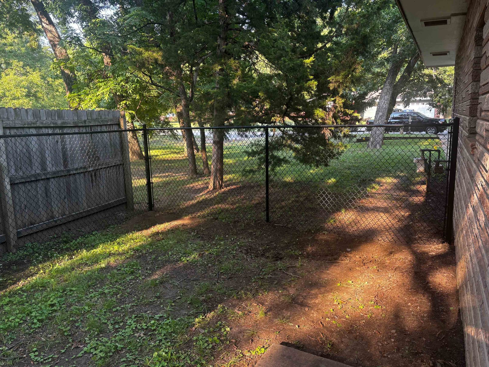 Backyard with chain-link fence, trees, and a dirt path beside a brick wall