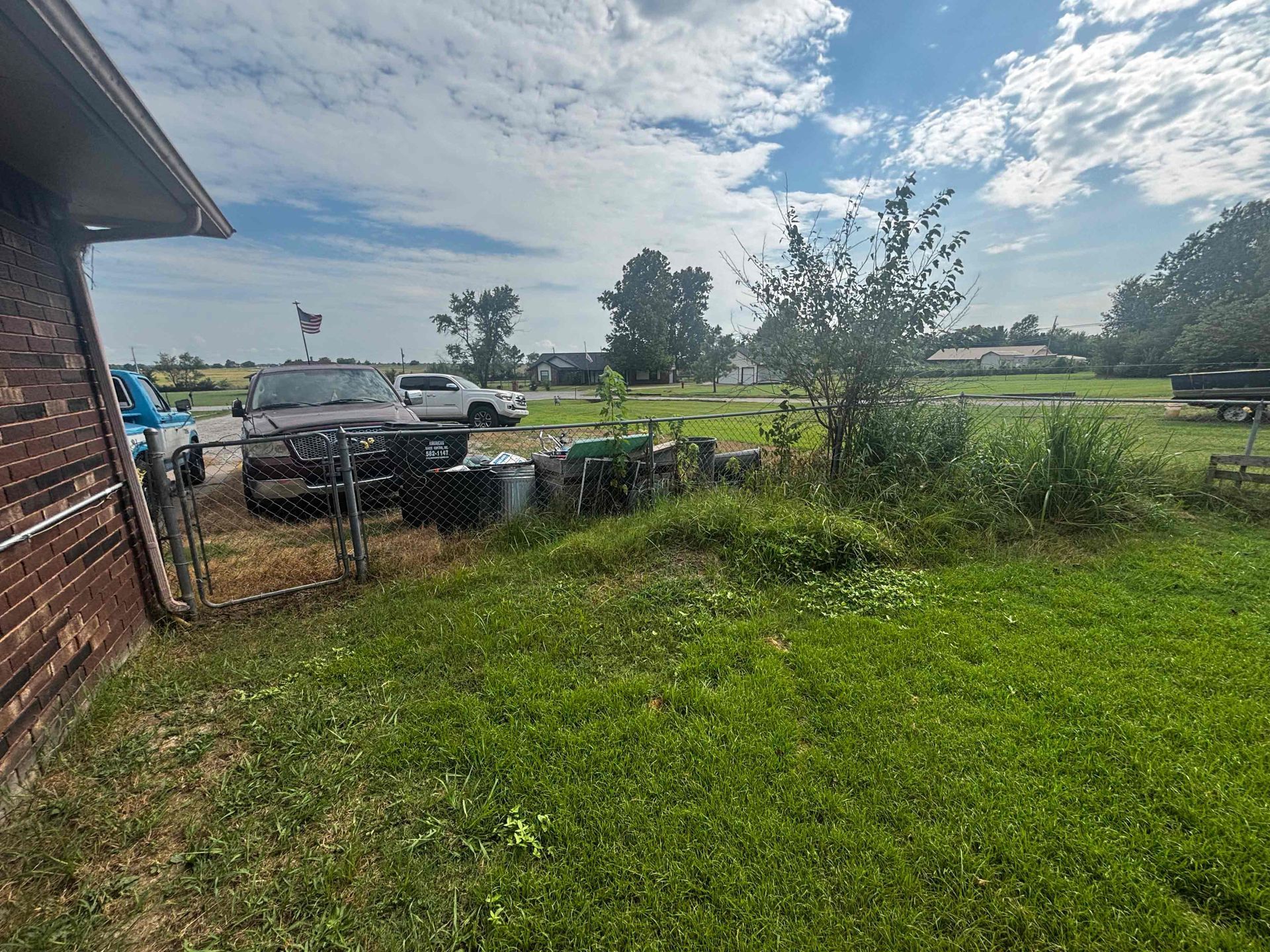 Rural yard beside a brick house, with parked vehicles, fence, and green field under a cloudy sky