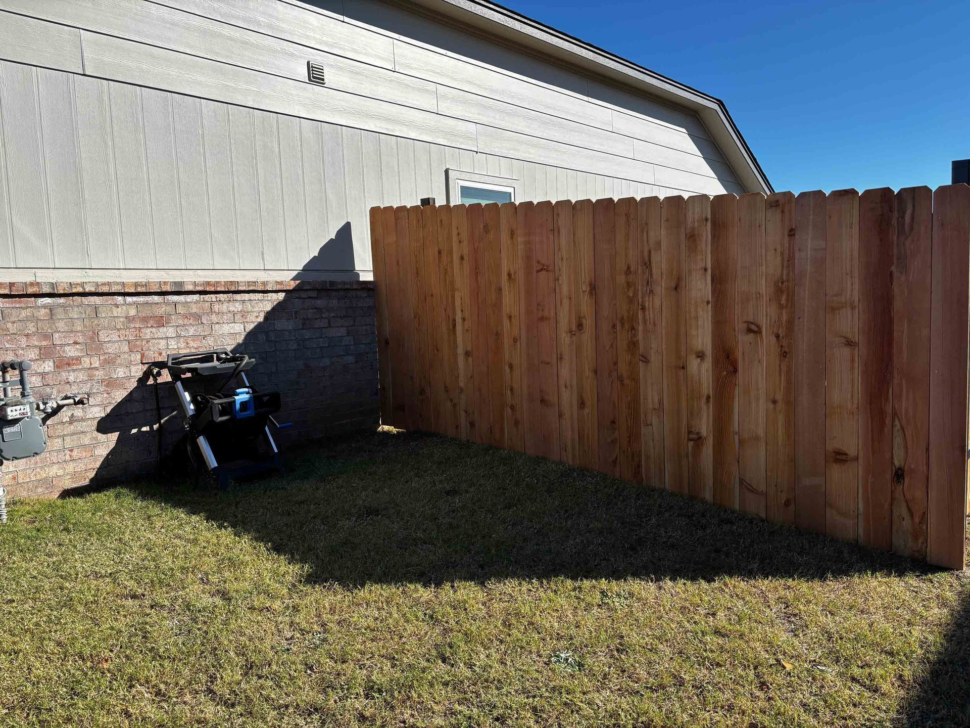 Wooden privacy fence beside a house, with a small black grill on a grassy backyard corner.
