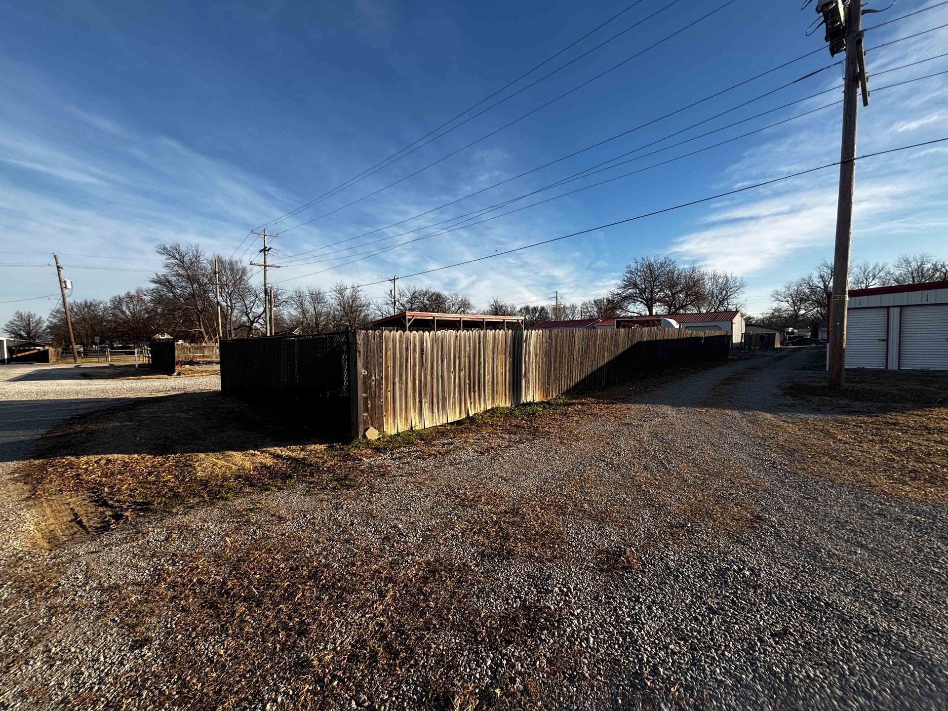 Gravel alley with a wooden fence and utility poles under a clear blue sky, beside houses and a shed.