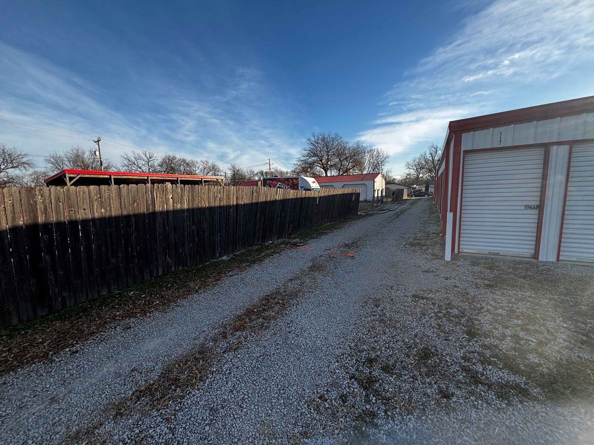 Gravel driveway beside a black fence and red storage building under a blue sky.