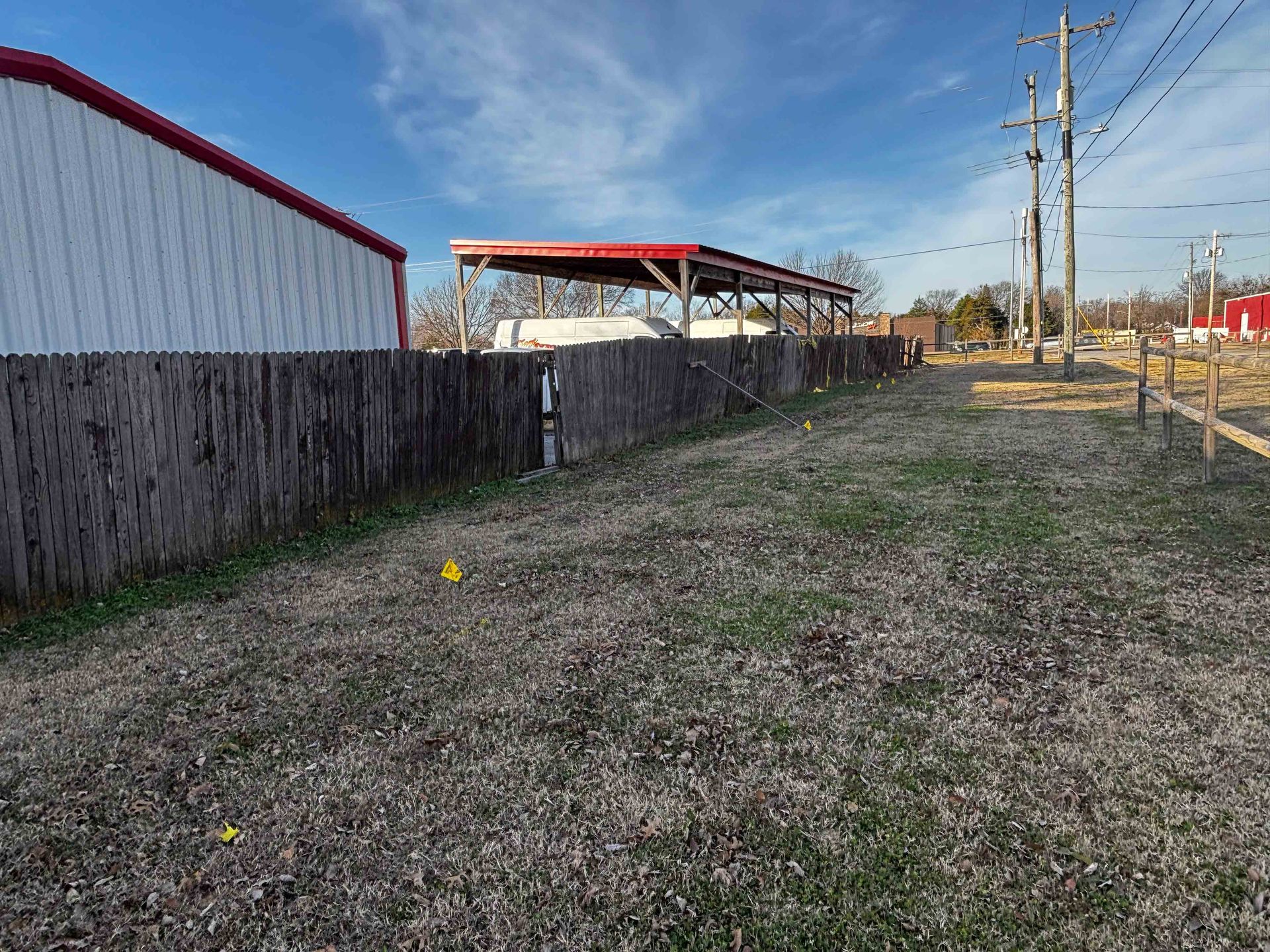 Grassy lot beside a fence and utility poles, with red-roofed buildings under a clear blue sky