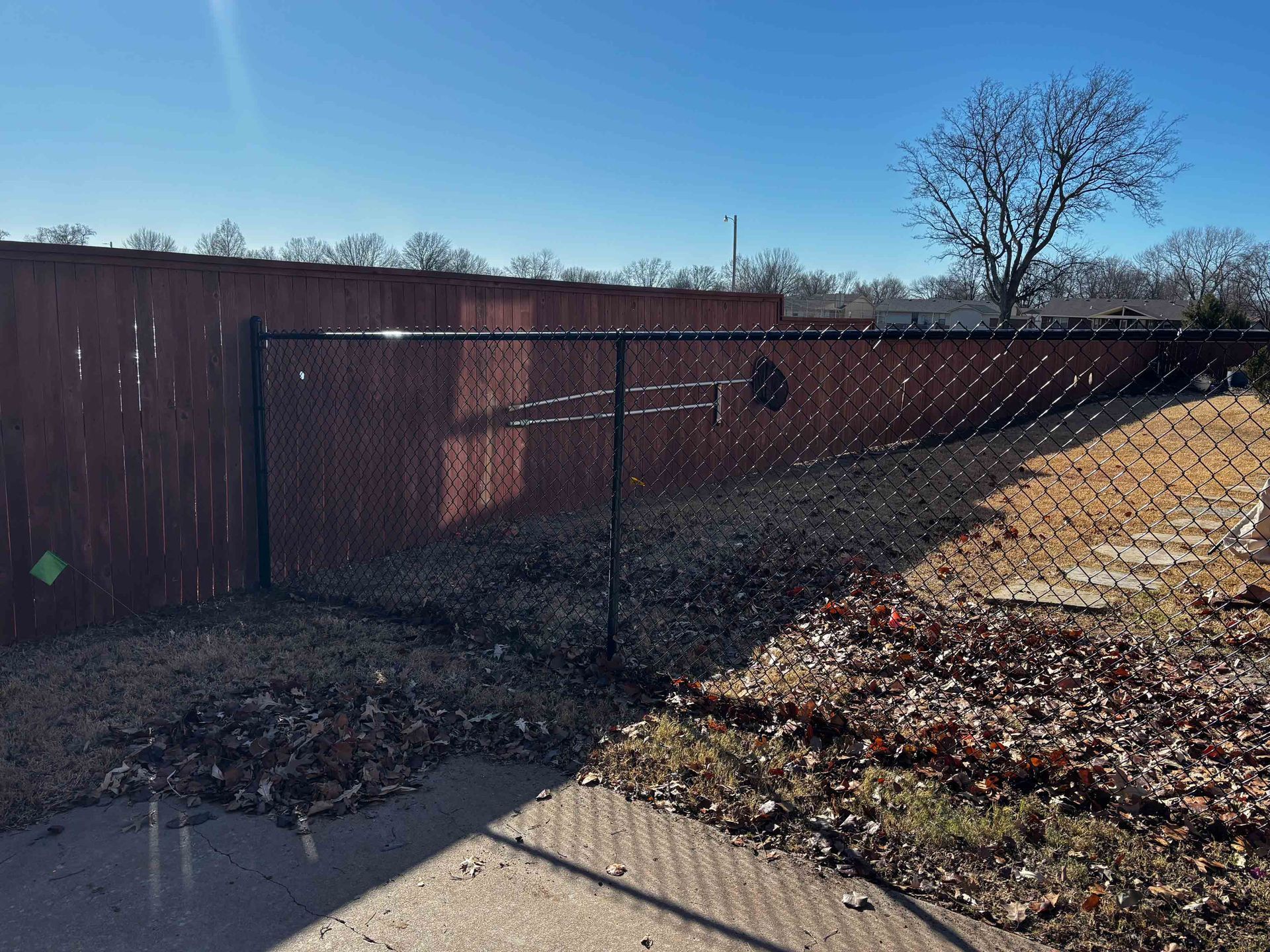 Chain-link fence beside a brown wooden fence under a clear blue sky, with dry leaves on the ground.