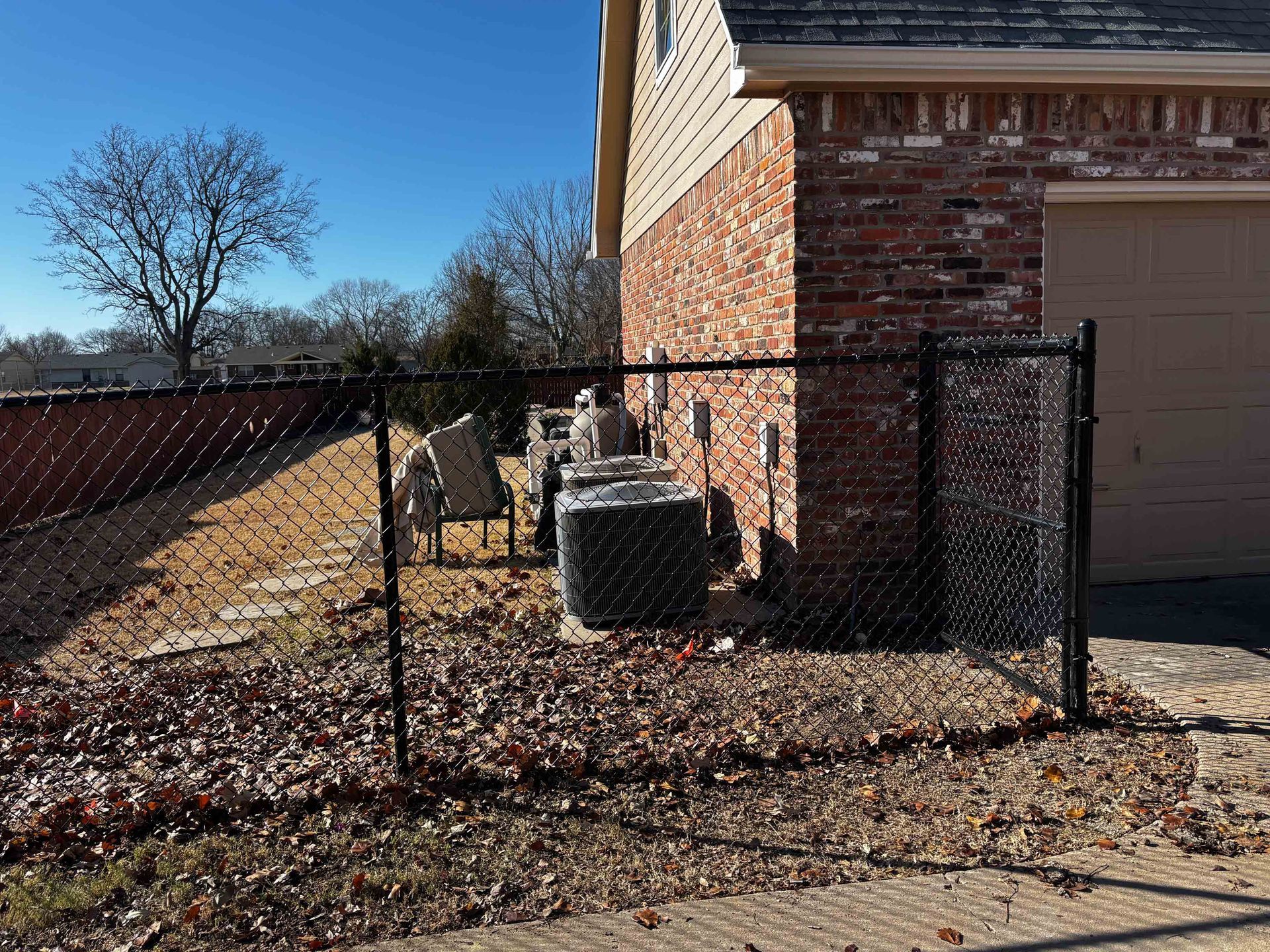 Backyard beside a brick house, with a fenced air-conditioning unit and leaf-covered ground