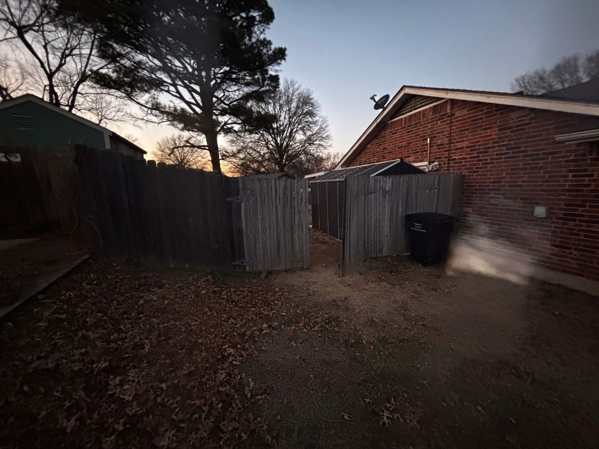 Backyard with wooden fence, shed, brick house, and bare trees at dusk