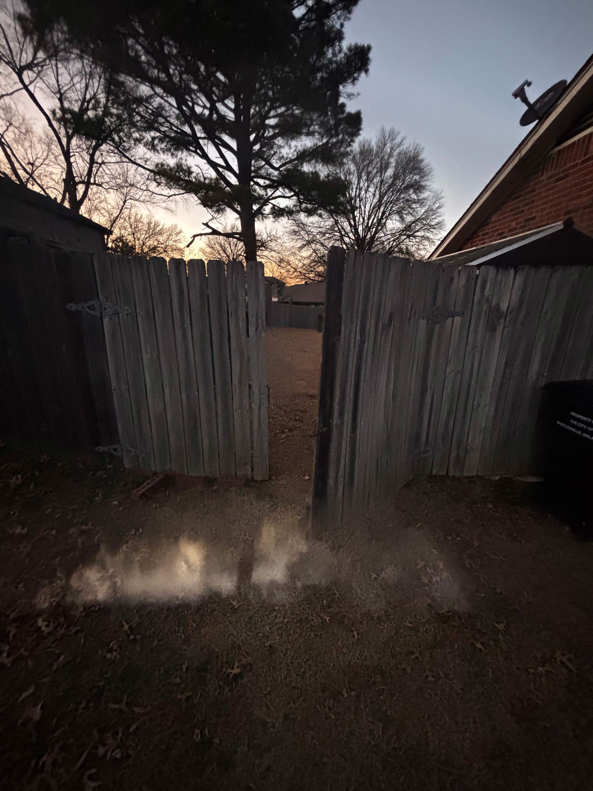 Dusk view of a muddy yard through open weathered wooden gates, with trees and a house roof silhouetted beyond.