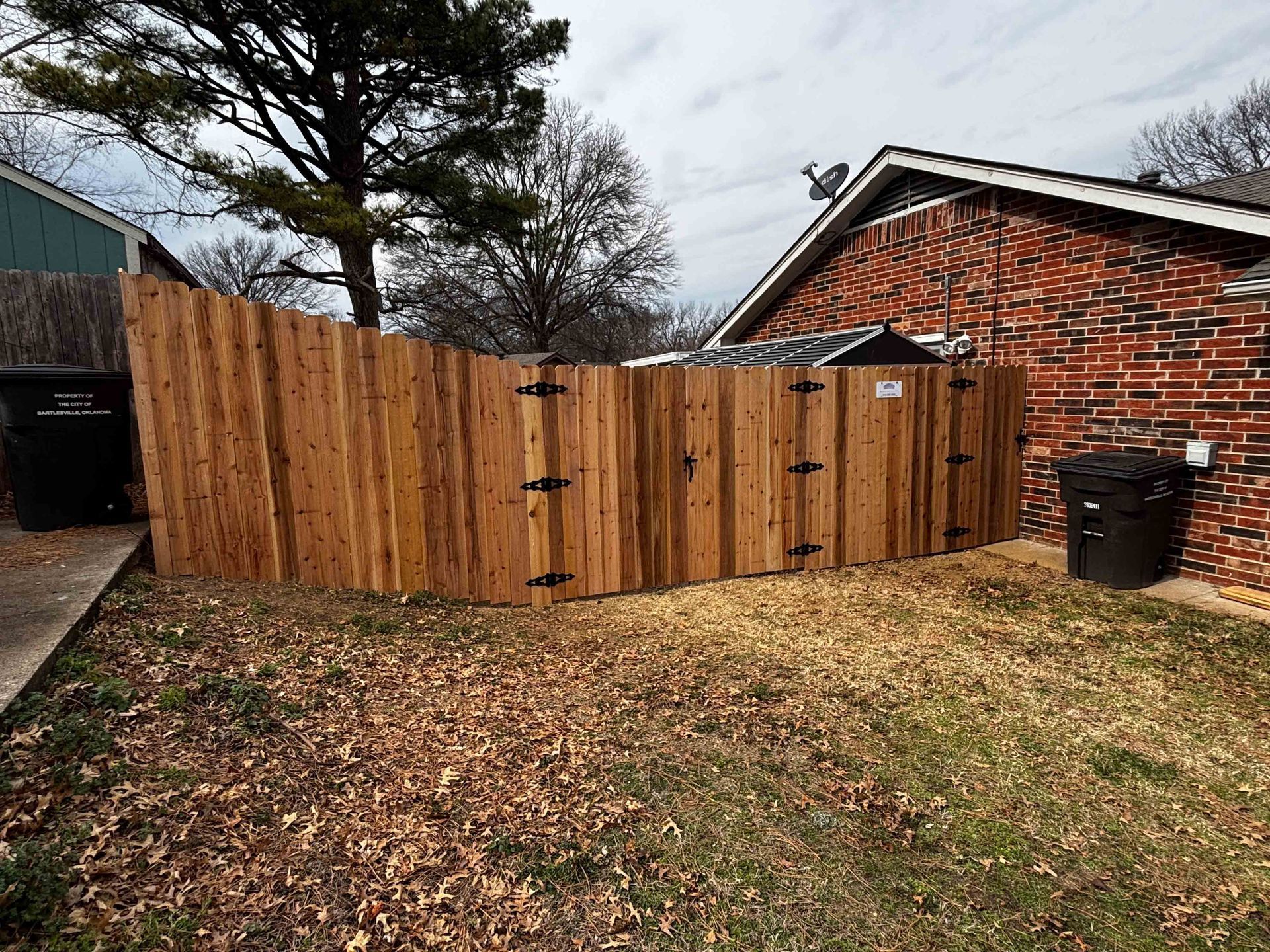Wooden fence gate beside a brick house, with a trash bin and leaf-covered yard