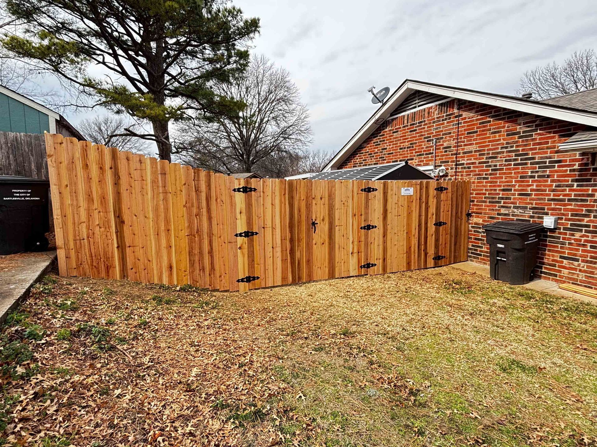 Wooden privacy fence in a leaf-covered yard beside a brick house and trash bins