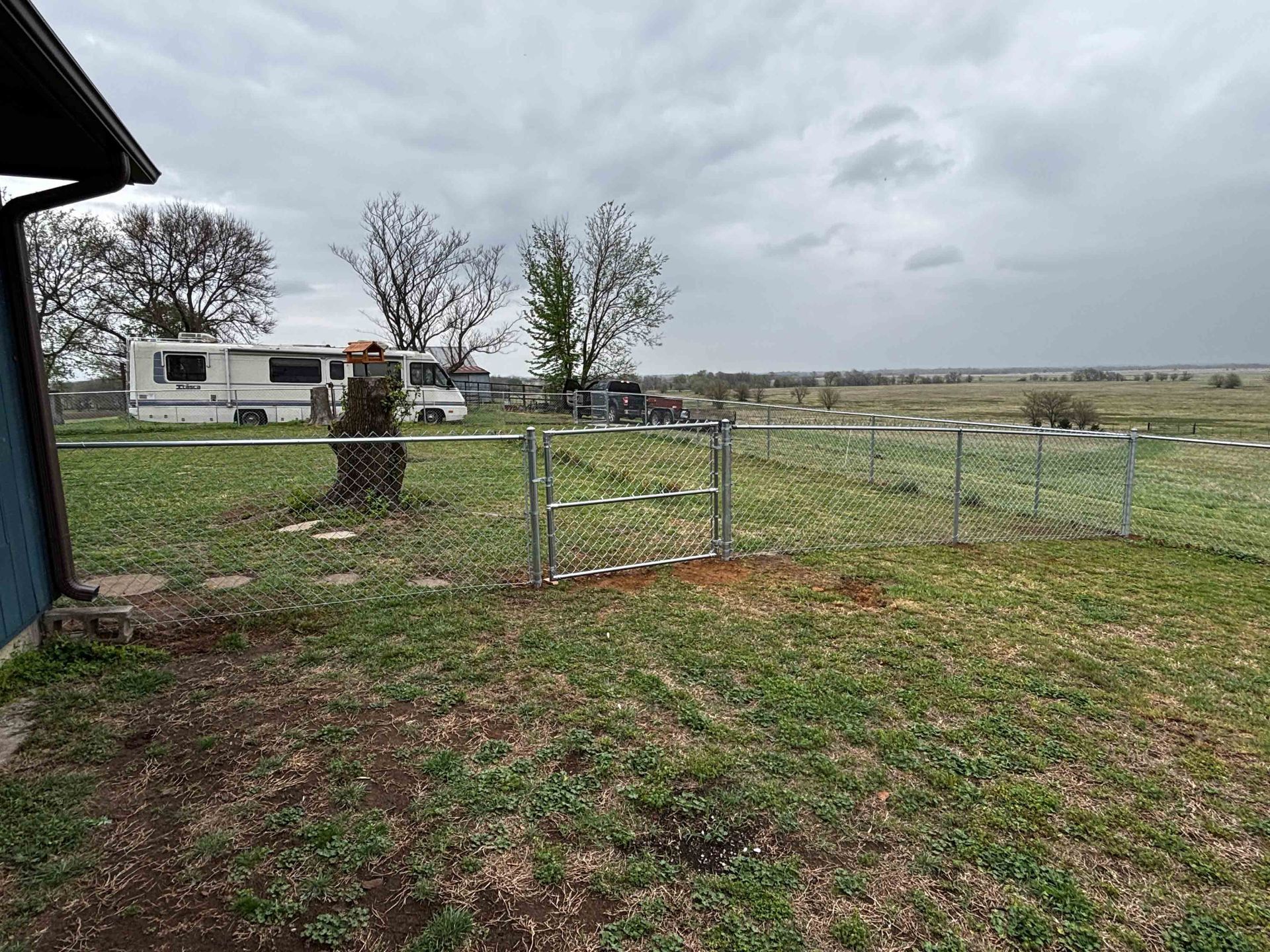 Rural yard with chain-link gate, white RV, tree stump, and cloudy sky over open fields