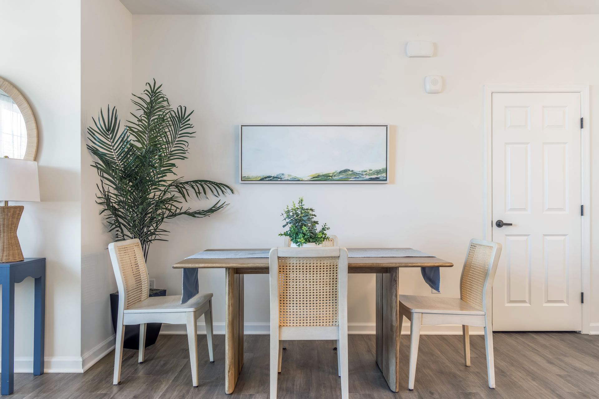 Dining room with table, chairs, art, and a large plant. White walls, gray floor.