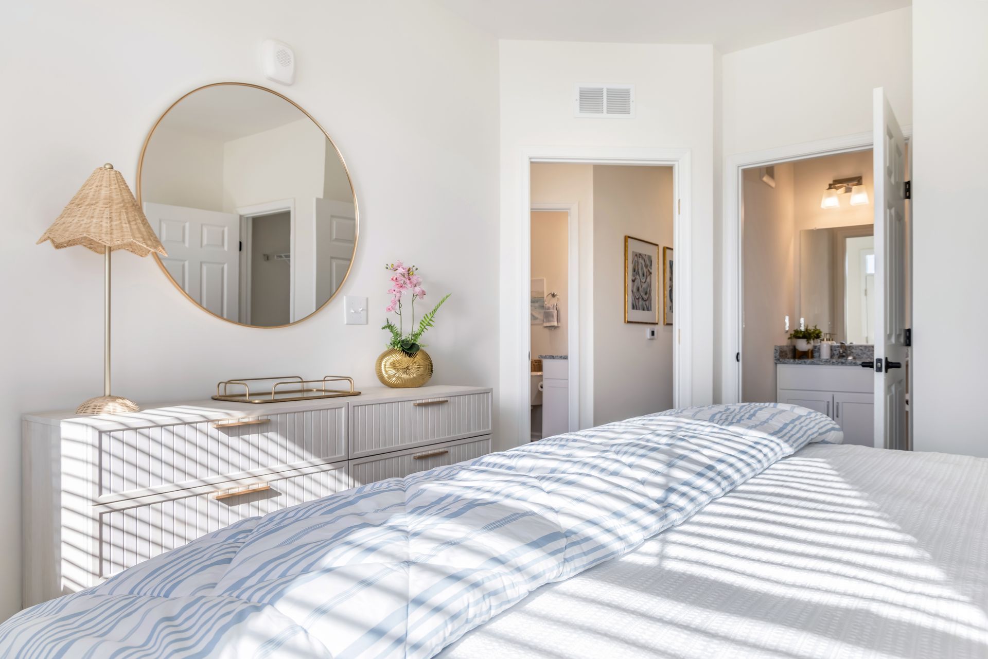 Bedroom with white walls, dresser, large round mirror, and a bed with blue and white striped bedding.