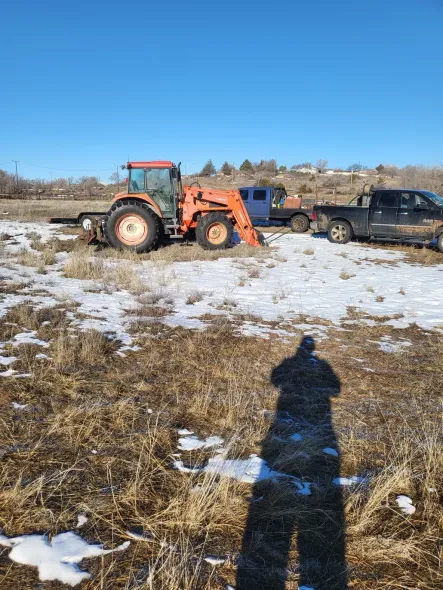 Orange tractor with loader on snowy field, shadow of person taking photo visible.