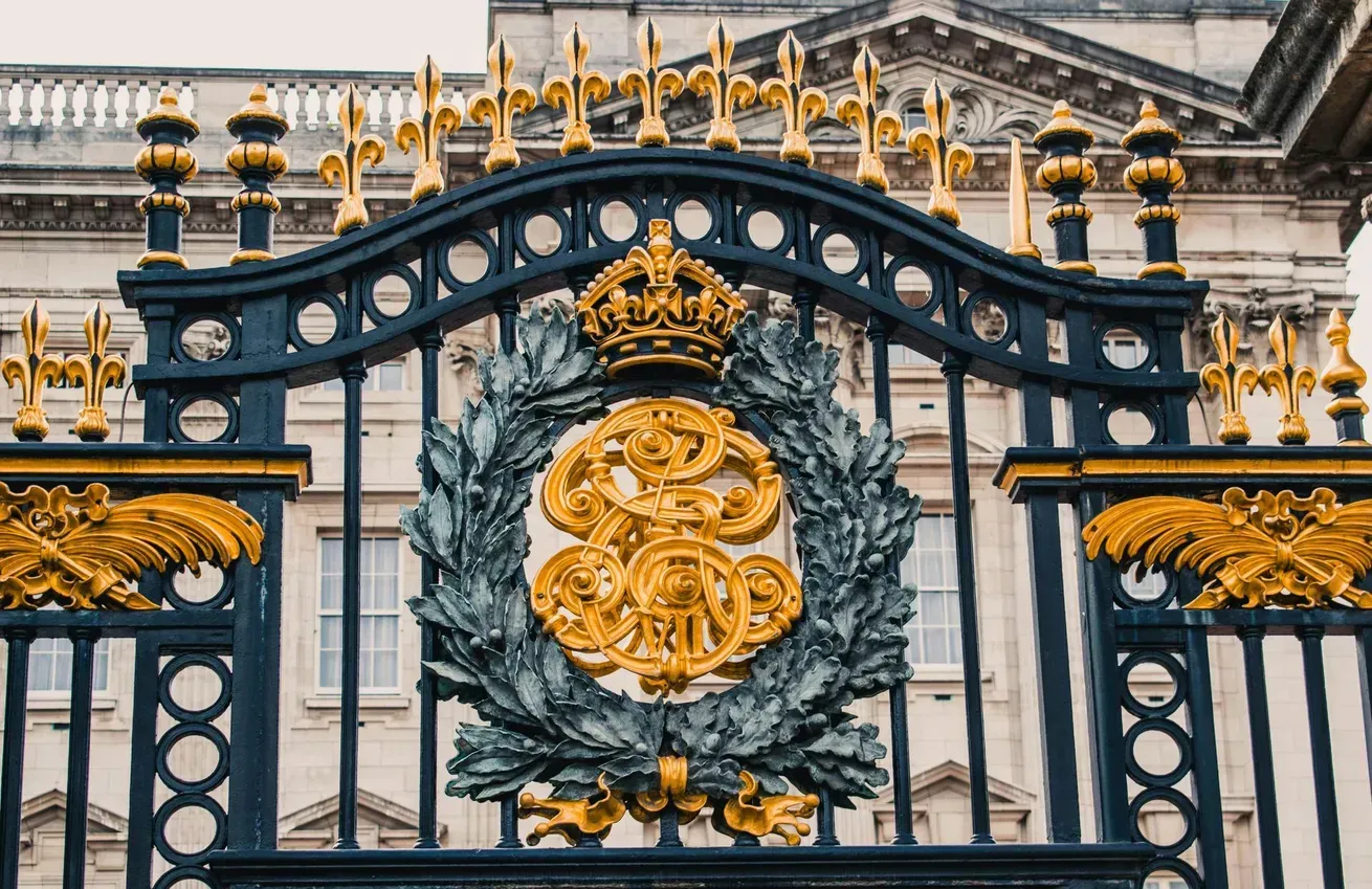 Ornate gold and blue iron gates with a royal crest; Buckingham Palace in the background.