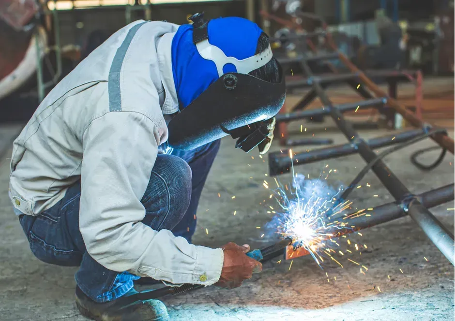 Welder in blue protective gear, squatting and welding metal with sparks in a workshop.