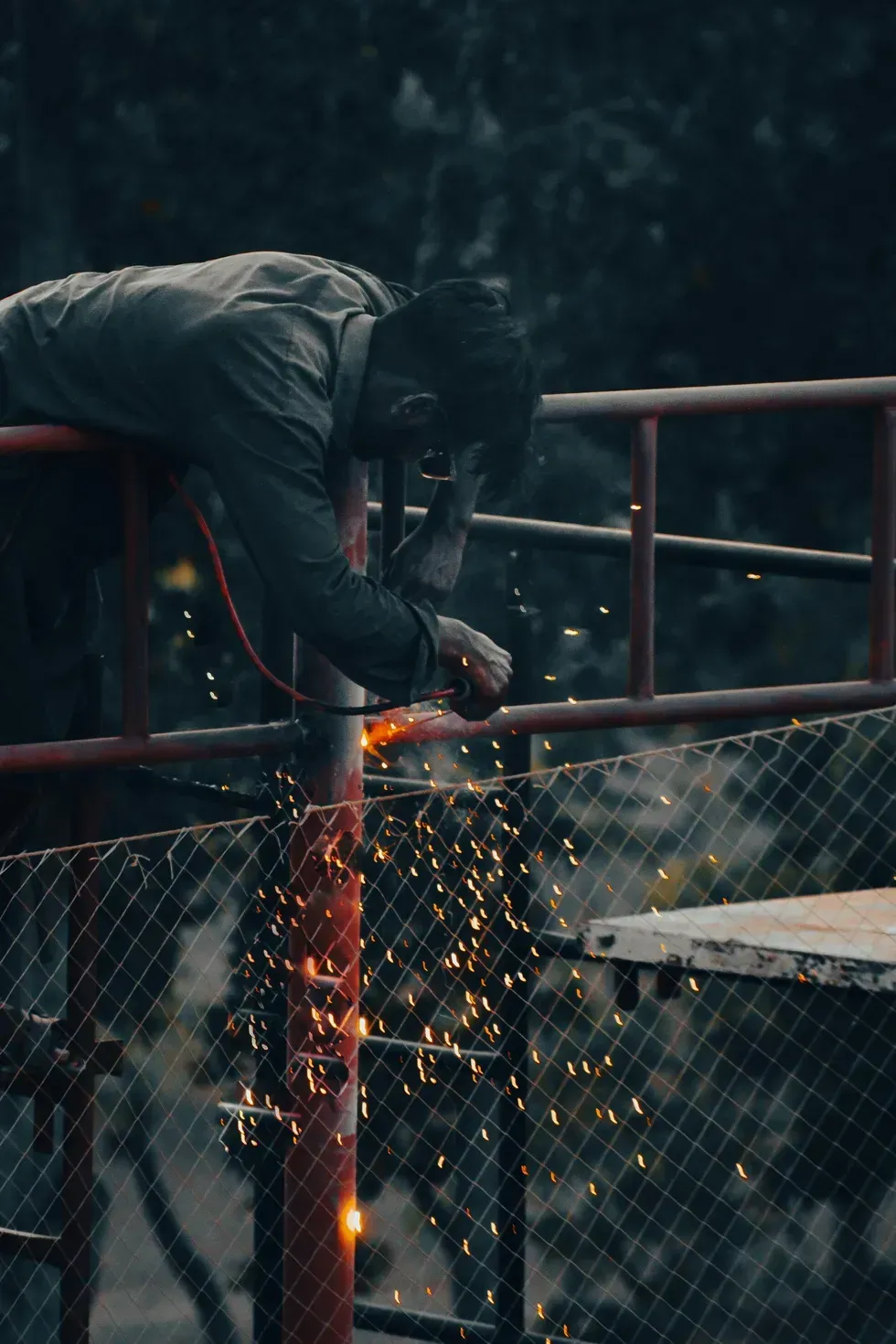 Person welds on a red railing, sparks flying, outdoors. Dark setting.