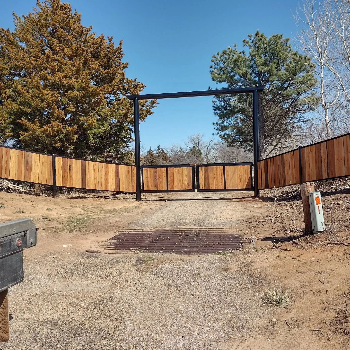 Gate and wooden fence on a dirt road, with trees and blue sky visible.