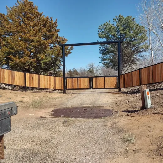 Gated driveway with wooden fence, surrounded by trees, under a blue sky.