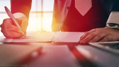 Person in a suit signing documents with a pen at a desk, near a window. Bright sunlight.