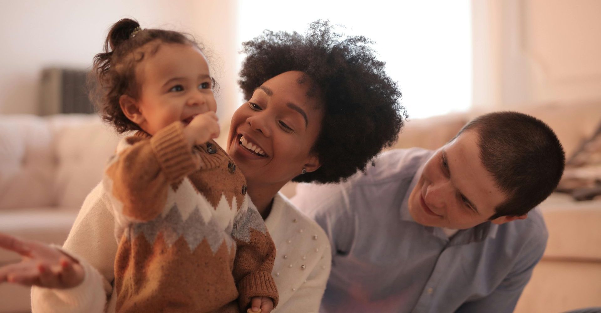 Smiling parents and a child indoors; the mother holds the baby, the father looks on, both happy.