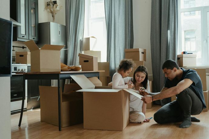 Family unpacking boxes in a new home. Boxes surround them. Mother, child, and father are looking inside a box.