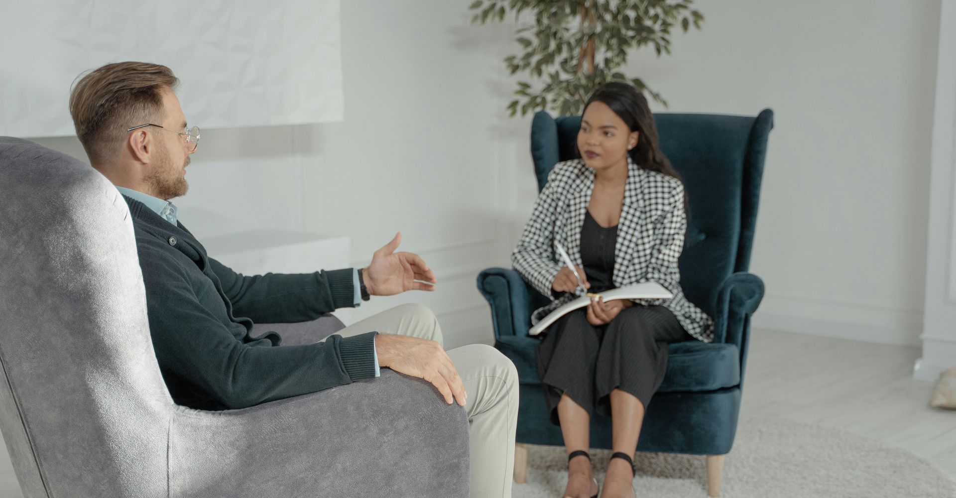A man in a chair talking to a woman in a blazer, sitting in an armchair with a notebook.
