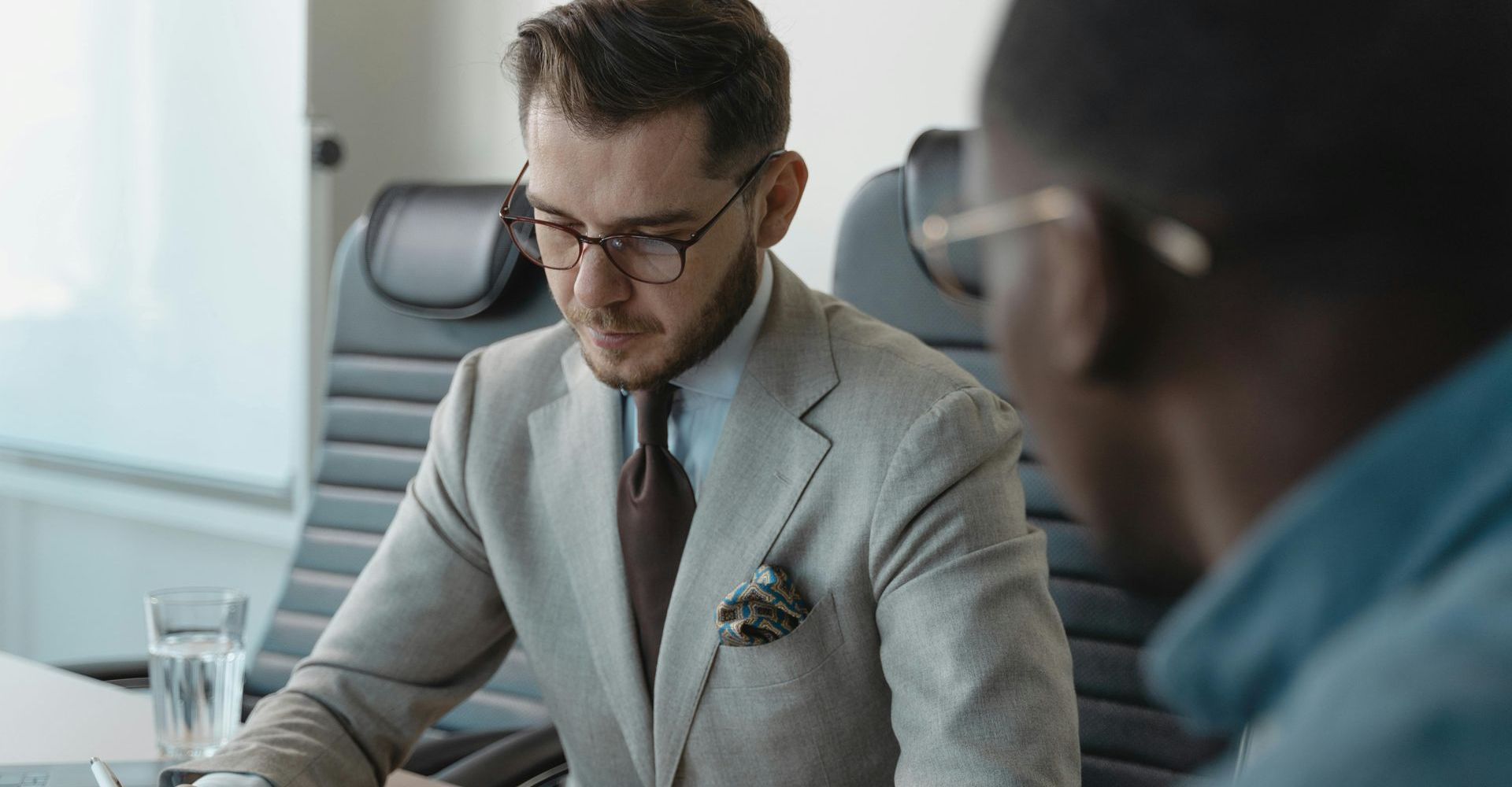 Two men in business attire at a table; one examines documents.