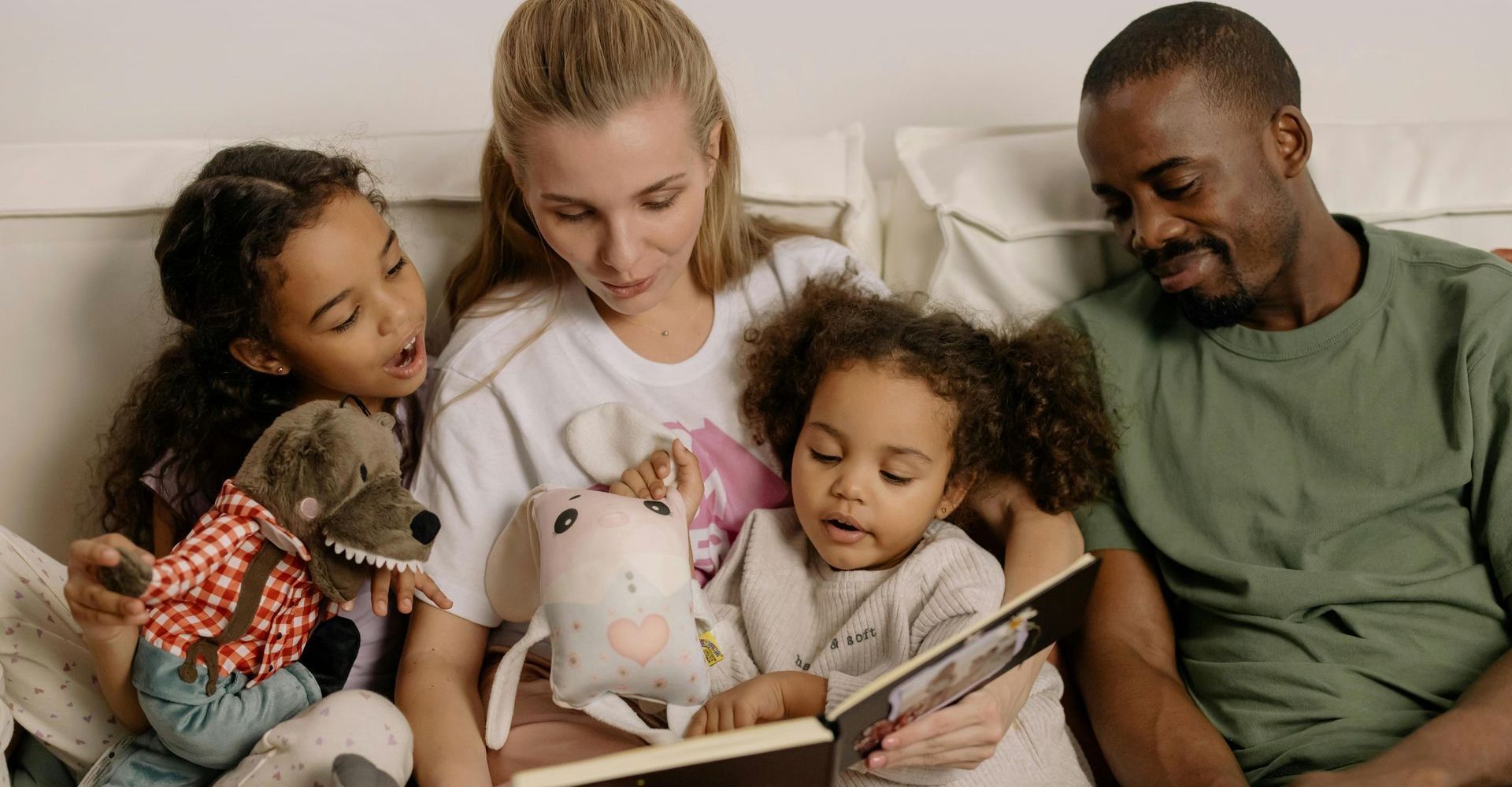 A diverse family reads a book together, smiling. Children hold stuffed animals.