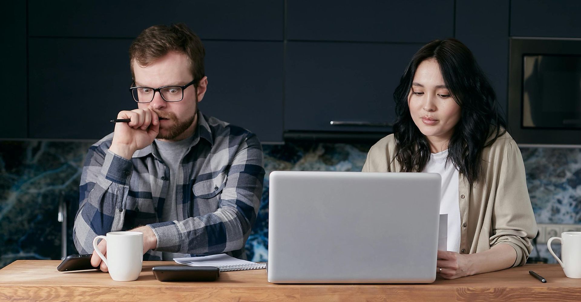 Couple working on laptop at a kitchen counter; man looks stressed, woman focused, with mugs and calculator nearby.