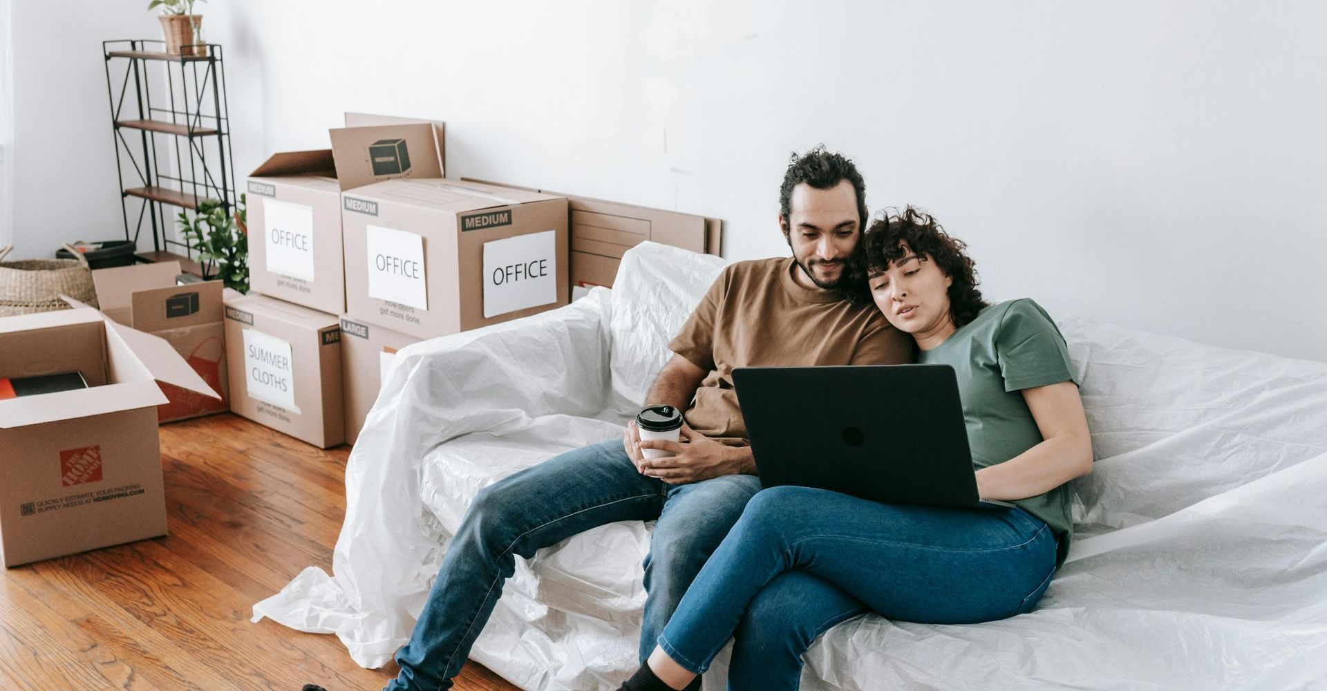 Couple on couch looking at laptop, surrounded by moving boxes in a bright room.