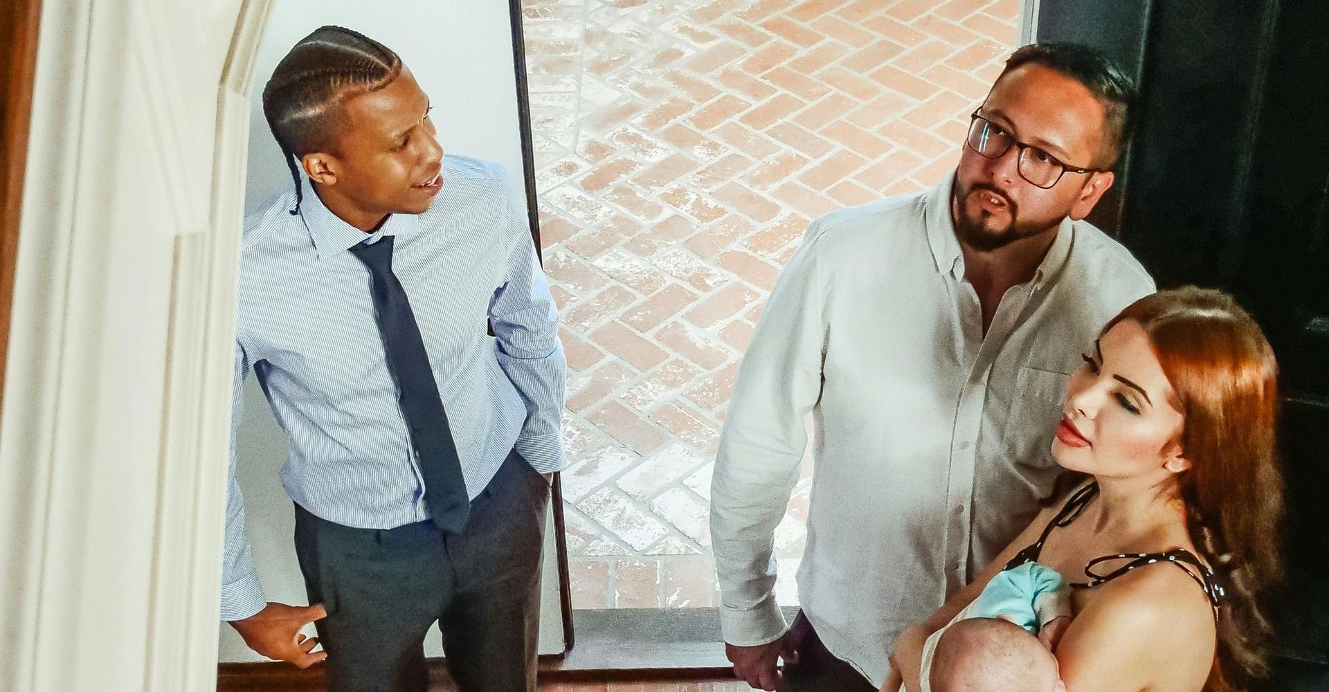 Man in a suit and tie peers at couple with a baby at a doorway.