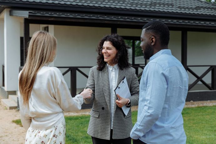 Real estate agent fist-bumps a woman in front of a house with a couple; agent smiling.