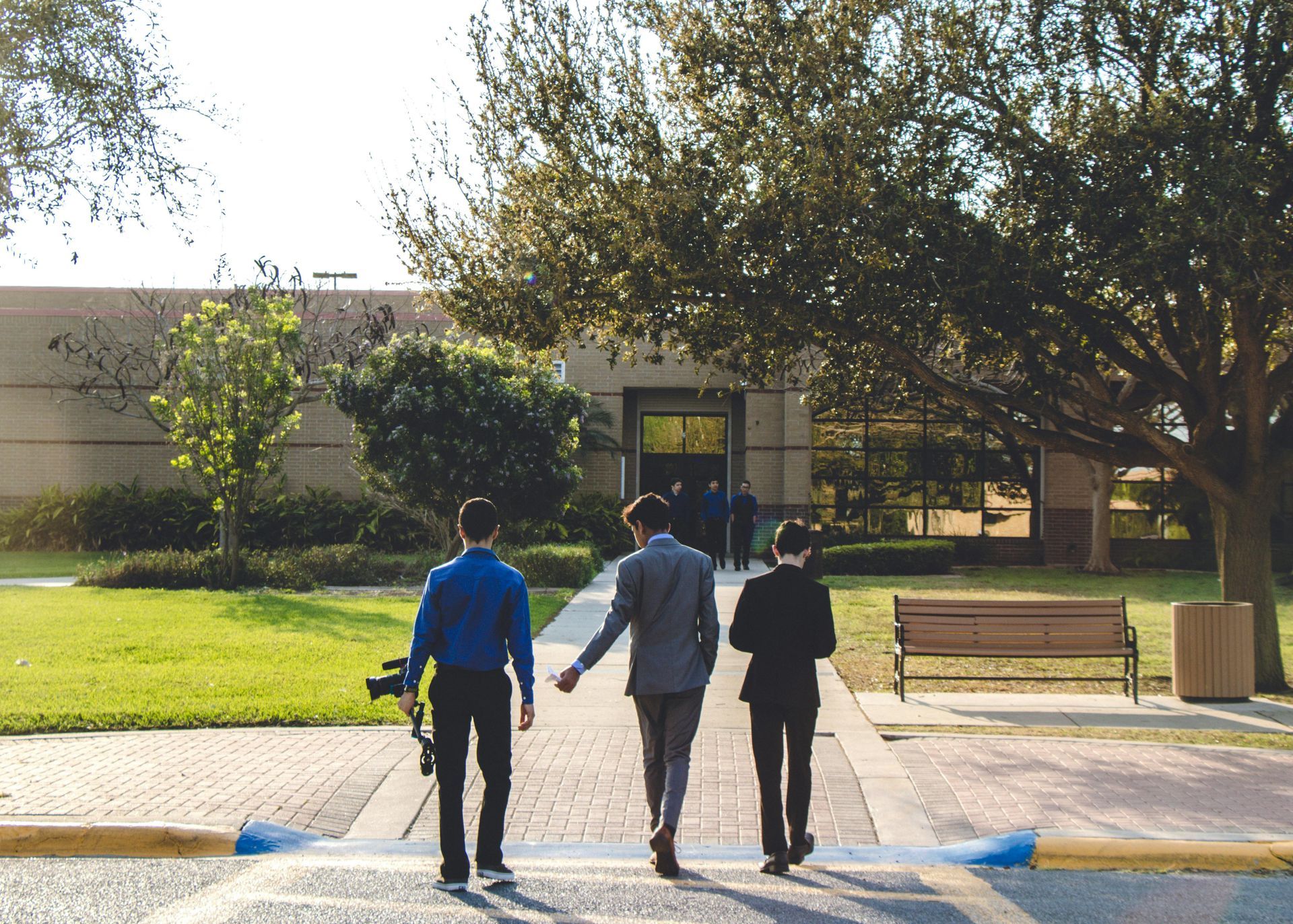 Three young men in suits walk toward a building entrance on a sunny day.