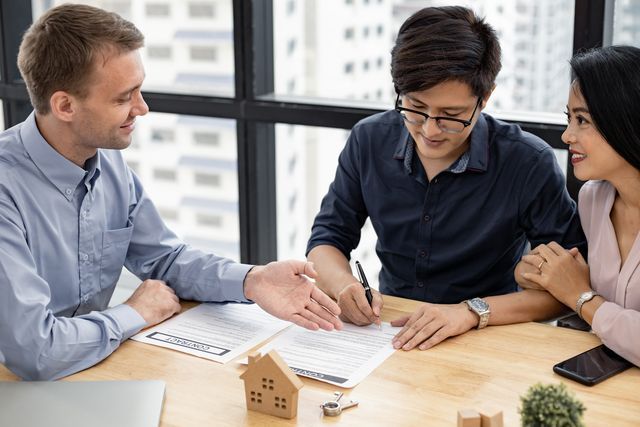 Real estate agent with couple signing a document at a table; small house model and keys present.