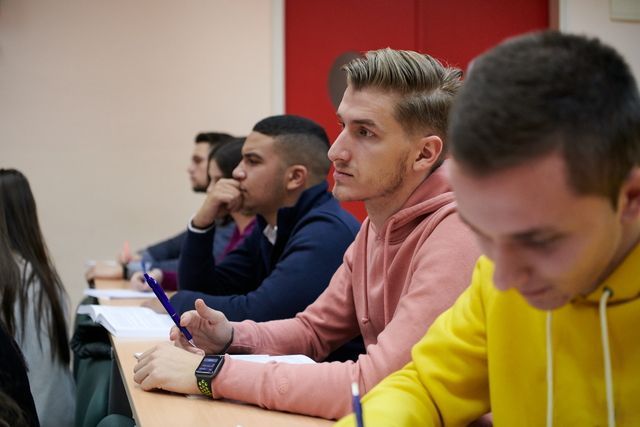 Students in a classroom, writing at desks. One man wears a pink hoodie and looks attentively ahead.