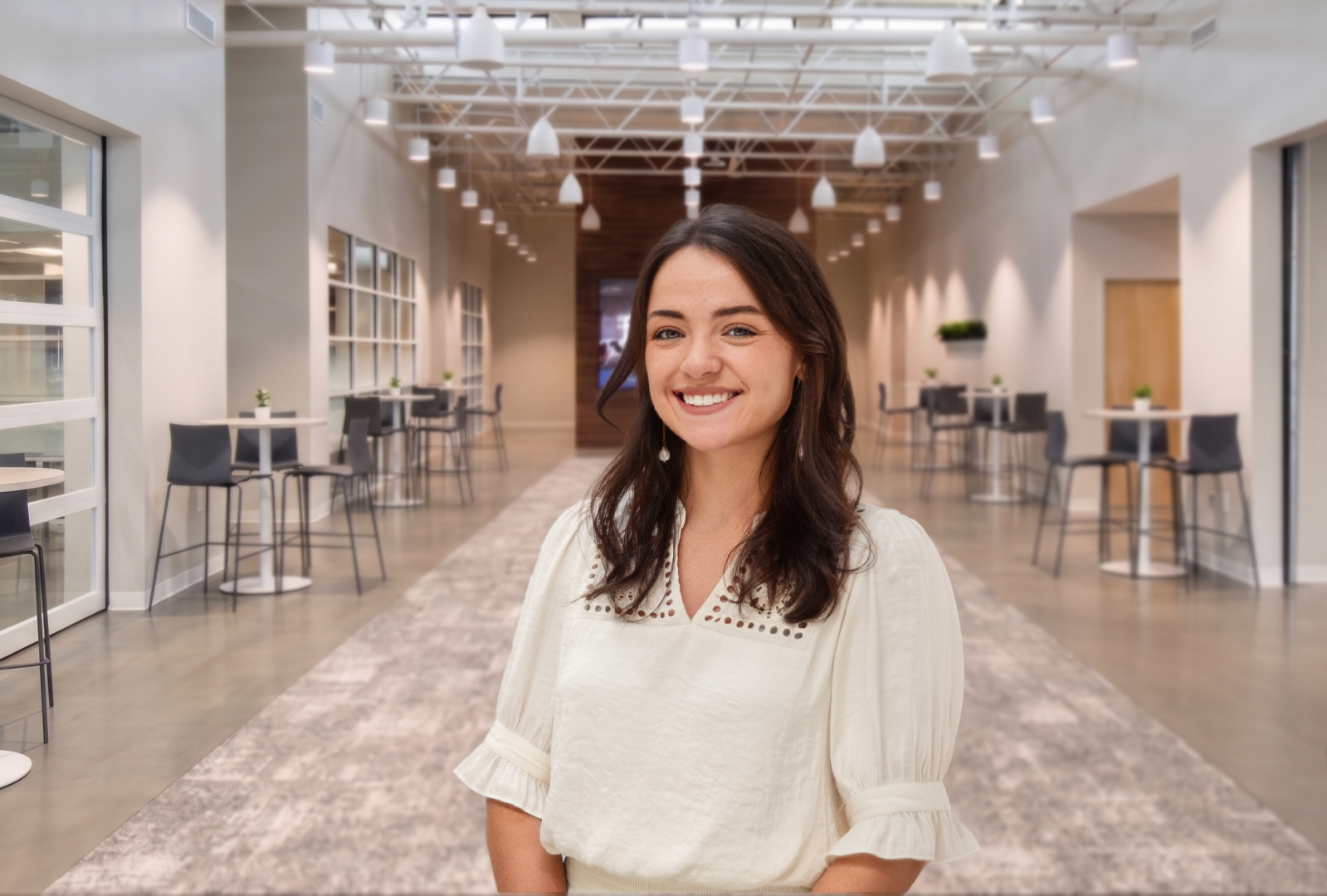 A smiling person in a cream-colored blouse stands in a bright, modern office hallway with tables and chairs.