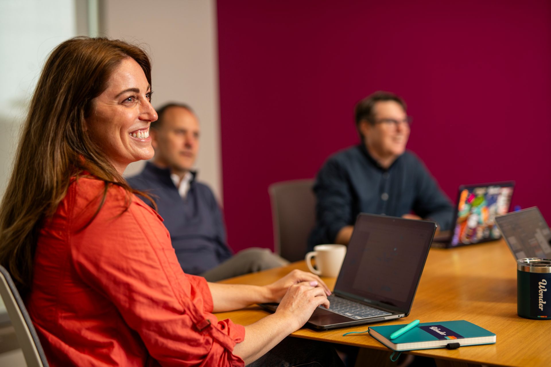 Woman in a red blouse using a laptop in a meeting room with two colleagues in the background