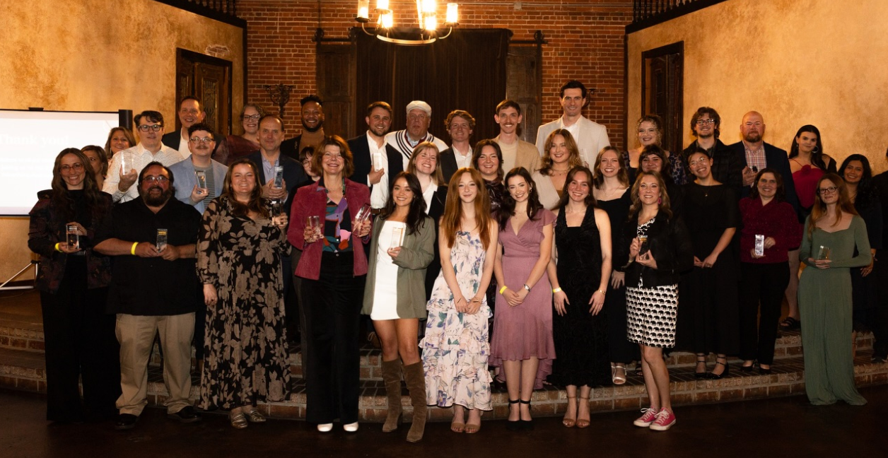 Large group posing indoors at a formal event, with brick walls and warm lighting.