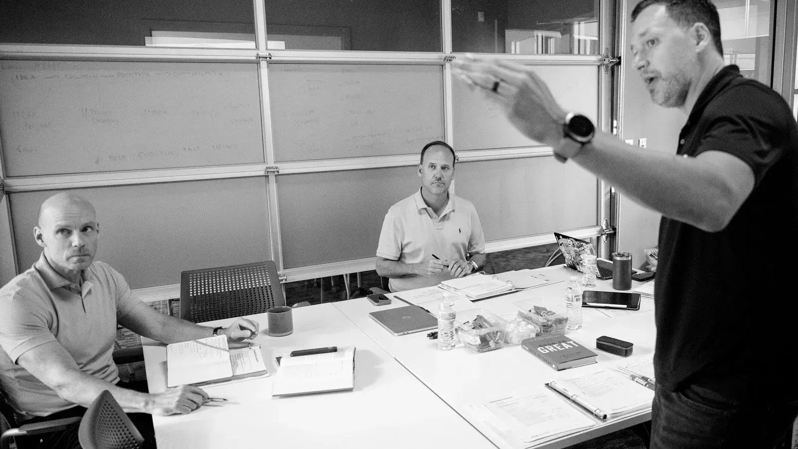 Three men in office, one gesturing, others seated at table with papers and objects.