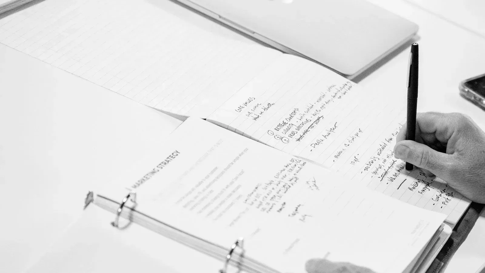 Person writing on lined paper in a binder, with a laptop in the background.